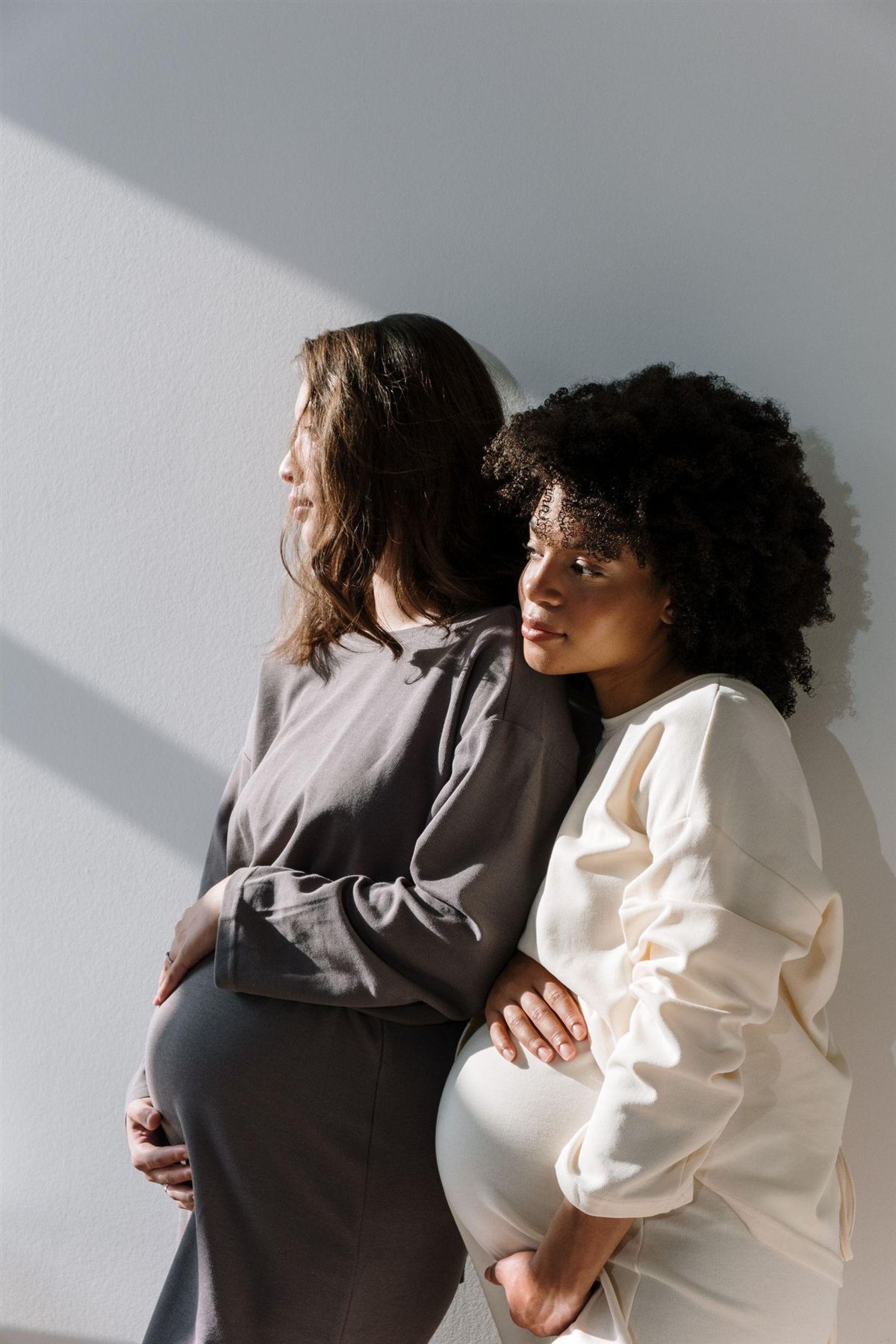 Two women standing together by a sunlit wall, one of them pregnant