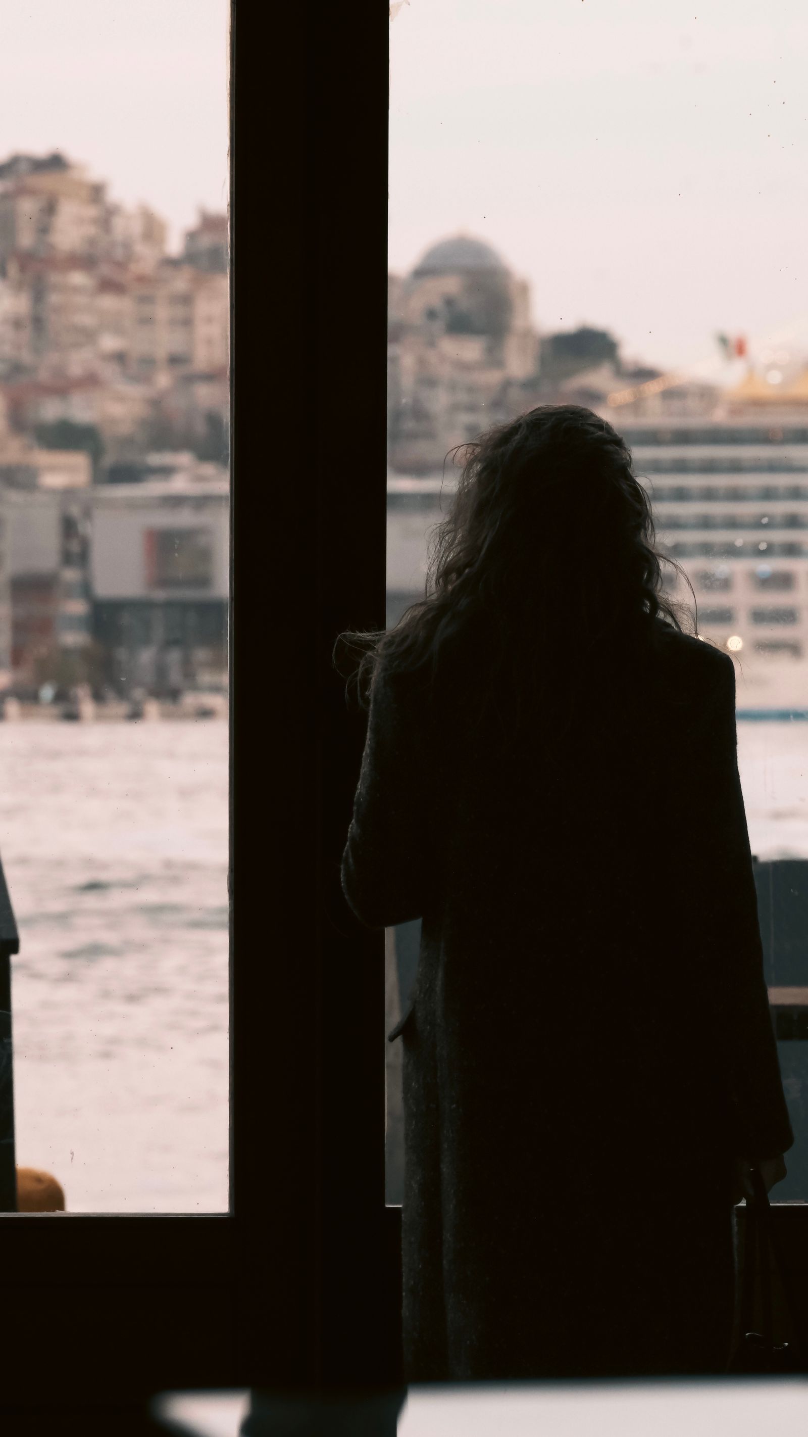 A woman standing quietly at a window looking out over the water